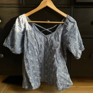 Blå broderad blus med snörning - Supersöt ljusblå blus med vita broderade blommor och hålmönster. Blusen har puffiga korta ärmar och snygg snörning i ryggen som ger en extra detalj. Perfekt för vår och sommar, och materialet känns lätt och luftigt.