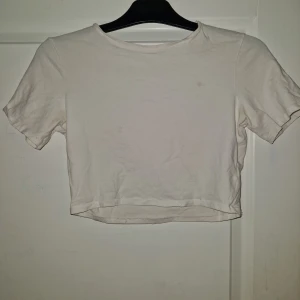 Vit croppad t-shirt i bomull - Enkel vit croppad t-shirt med rund hals och korta ärmar. Tillverkad i mjuk bomull, perfekt för en avslappnad stil. Passar snyggt till både jeans och kjol. (Liten fläck tyvärr)