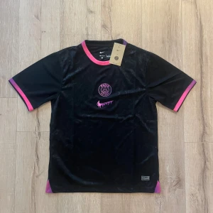 PSG x Nike svart fotbollströja M - Snygg svart Paris Saint-Germain fotbollströja från Nike med rosa och lila detaljer på krage och ärmslut. Tröjan har PSG-logga och ett unikt rosa Nike-märke på bröstet. Tillverkad i Dri-FIT polyester med subtilt blommönster i tyget. Perfekt för dig som gillar streetwear och fotboll.