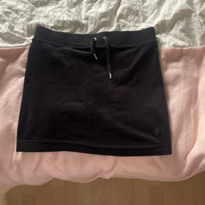 Svart kortkjol från Juicy Couture - Svart kortkjol från Juicy Couture med snörning i midjan och diskret logga broderad längst ner. Kjolen är rak i modellen och gjord i mjukt bomullsmaterial, perfekt för en chill och sportig vibe. Metalländarna på snöret har Juicy Couture-gravyr för extra detalj.