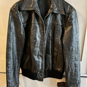 Svart skinnjacka från danska Bzr - hög kvalitet  - Snygg svart skinnjacka i klassisk modell med bred krage och dragkedja framtill. Jackan är från det danska märket Bzr (underlinje till Bruuns Bazar) och i mycket hög kvalitet. I buffelskinn och obetydligt använd. Jackan har ribbade muddar vid midja och ärmslut, samt två innerfickor. Perfekt för dig som vill ha en tidlös och cool look. Bredd cirka 53 cm oc äh längd cirka 62 cm (bak), Ärmarna är ungefär 66 cm långa (från axeln). 