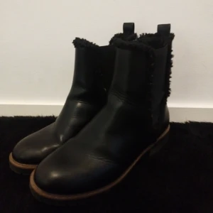 Svarta boots i skinn med foder - Säljer ett par svarta boots i skinn med grov sula och mjukt foder längs kanten. De har rund tå, bruna detaljer vid sulan och dragflikar upptill. Perfekta för kalla dagar och ger en clean look till vilken outfit som helst.
