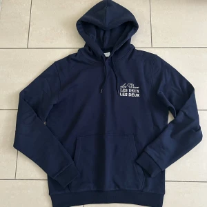 Mörkblå hoodie från Les Deux - Snygg mörkblå hoodie från Les Deux med vit logga tryckt på bröstet. Klassisk känguruficka, huva med snörning och ribbade muddar. Perfekt för chill dagar och enkel att matcha med jeans. Originalpris runt 1600kr och pris kan diskuteras!