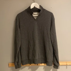 Mörkgrå tröja från H&M med krage - Snygg mörkgrå tröja från H&M med ribbad krage och v-detalj vid halsen. Tröjan har långärmade ärmar och är tillverkad i mjuk bomullsmix som är skön att ha på sig. Perfekt för dig som gillar enkel och stilren design.