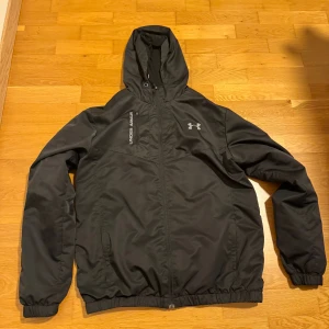 Svart vindjacka från Under Armour XL - Svart vindjacka från Under Armour i storlek XL, Men passar som L. Jackan har huva, dragkedja framtill, två sidofickor och elastiska muddar. Materialet är lätt och syntetiskt, perfekt för blåsiga dagar. Snygg logga på bröstet och diskret branding längs axeln.