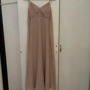 Beige långklänning från ASOS DESIGN - Säljer en elegant beige långklänning från ASOS DESIGN Petite med tunna axelband och v-ringad överdel. Klänningen har ett flytande, lätt tyg och dragkedja i ryggen. Perfekt för dig som gillar stilrena och romantiska looks.