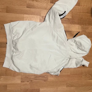 Vit hoodie från EA7 Emporio Armani - Snygg vit hoodie från EA7 Emporio Armani med svart dragkedja och svart innerfoder i huvan. EA7-logga på bröstet och tryck på ena axeln. Hoodien har långa ärmar, huva med snörning och två fickor framtill. Perfekt för en chill och sportig look.