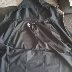 Svart vindjacka från Under Armour XL - Svart vindjacka från Under Armour i storlek XL. Jackan har huva, dragkedja framtill och är tillverkad i lätt polyester med meshfoder. Perfekt för aktiva dagar och sportiga outfits. Diskret logga på bröstet och sportig passform.