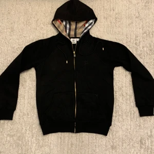 Svart Burberry hoodie - Svart hoodie från Burberry i M  men passar i S bättre. Tröjan har broderad Burberry-logga på bröstet, snörning vid huvan och två fickor framtill. Perfekt för dig som gillar stilrena och lyxiga detaljer.