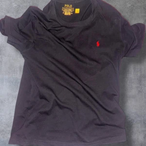 Svart t-shirt från Polo Ralph Lauren - Svart t-shirt från Polo Ralph Lauren med klassisk röd broderad logga på bröstet. T-shirten har rund halsringning och korta ärmar. Tillverkad i mjuk bomull för en skön känsla. Perfekt för en clean och stilren look.