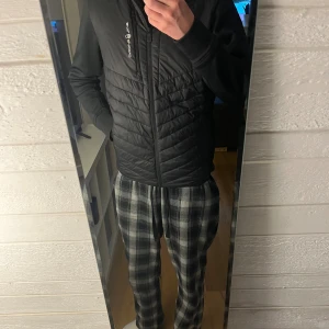 Svart quiltad pufferjacka från Denzling - Svart pufferjacka från sail racing med huva och dragkedja. Jackan har quiltad framsida och baksida, släta ärmar och en sportig look. Perfekt för kyliga dagar och enkel att matcha med allt. Snygg logga på bröstet och huvan.