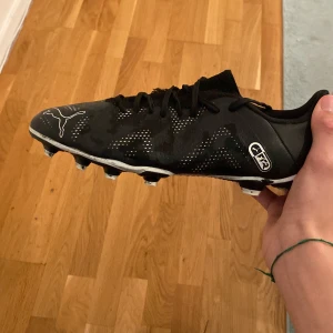 Svarta fotbollsskor från Puma - Säljer ett par svarta fotbollsskor från Puma med vita detaljer och snörning. Skorna har en låg profil och mönstrad ovandel med subtilt kamouflagemönster. Sulan har fasta dobbar för bra grepp på planen. Perfekt för dig som vill ha stil och funktion på matchen.