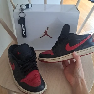 Nike Air Jordan 1 Low svart/röd - Nike Air Jordan 1 Low sneakers i svart och rött skinn med klassisk röd swoosh och Jumpman-logga på plösen. Låg modell med snörning och rund tå, samt röd yttersula. Perfekt för dig som vill ha en ikonisk streetwear-look. Ny pris 1600 kr! Skick 7/10!