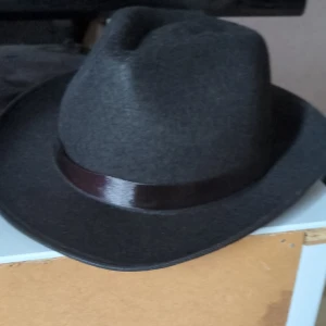 Svart klassisk hatt med band - Stilren svart hatt med brett brätte och ett mörkt, glansigt band runt kronan. Perfekt för dig som vill ha en tidlös och cool accessoar. Hatten har en snygg formad topp och är tillverkad i mjukt material som ger en elegant look. Helt ny och har bara använts en gång