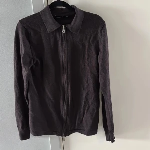 J lindberberg tröja - Snygg mörkbrun kofta från J.Lindeberg med hel dragkedja framtill och klassisk krage. Långärmad modell i mjukt stickat material, perfekt för lager-på-lager-stil. Passar dig som gillar stilrena och enkla plagg med en twist.