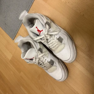 Nike Air Jordan 4 vita sneakers - Nike Air Jordan 4 sneakers i vitt med grå och röda detaljer. Skorna har snörning, meshpaneler på sidorna och en synlig Air-enhet i sulan. Ikonisk Jumpman-logga på plösen och hälen. Tillverkade i skinn och syntetmaterial med platt sula och rund tå. 