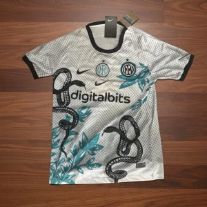 Inter Milan fotbollströja - Snygg vit Inter Milan fotbollströja från Nike med svart och turkost ormmönster och digitalbits-logga på bröstet. Tröjan har korta ärmar, svart krage och detaljerade grafiska inslag. Perfekt för dig som gillar unik design och fotboll.