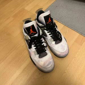 Nike Air Jordan 4 sneakers vit/grå - Nike Air Jordan 4 sneakers med vit och grå ovandel i syntetmaterial och mesh, svarta snören och detaljer i plast. Ikonisk Jumpman-logga i orange på plösen och under sulan. Snygg retrostil med rund tå och platt sula, perfekt för dig som gillar streetwear.