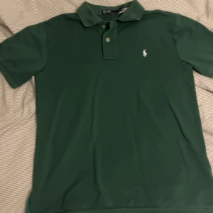 Grön pikétröja från Ralph Lauren - Snygg grön pikétröja från Ralph Lauren, modell The Earth Polo. Klassisk krage, korta ärmar och två knappar framtill. Ikoniska vita Polo-loggan broderad på bröstet. Perfekt för dig som gillar stilrena och tidlösa plagg.