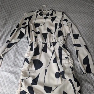 Vit och svart mönstrad kortklänning - Snygg kortklänning med lång ärm i vit och svart abstrakt mönster. Klänningen har en hög hals och är tillverkad i ett slätt, mjukt material som ger en cool och modern vibe. Passformen är normal och midjan är lätt markerad med rynkade detaljer på sidorna.