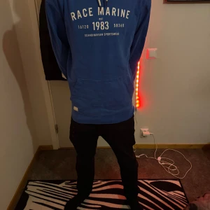 Blå hoodie från Race Marine M - Säljer en blå hoodie från Race Marine i storlek M. Tröjan har vit text på ryggen, huva med snören och en stor magficka. Perfekt för dig som gillar sportig och avslappnad stil. Mjuk insida och klassisk passform.