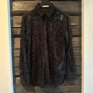 Svart spetsblus från Gestuz - Svart långärmad skjorta från Gestuz i transparent spets med blommigt mönster. Klassisk krage, knäppning framtill och breda manschetter med knappar. Perfekt för dig som vill sticka ut med en edgy och elegant look. Aldrig använd. Något stor i storleken. 