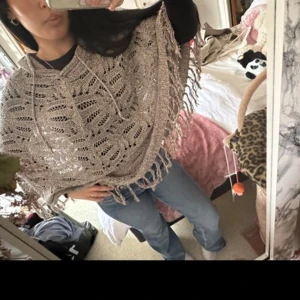 Beige stickad poncho  med fransar och huva - Säljer en beige stickad poncho  med snyggt hålmönster, fransar längs nederkanten och en stor huva. Västen har snörning vid halsen och är loose fit, perfekt att slänga över en t-shirt eller långärmad. Materialet är mjukt och ger en chill vibe.