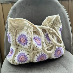 Beige virkad axelväska med blommor - Superfin handgjord axelväska i virkat material med beige botten och virkade blommor i lila, rosa och vitt. Väskan har rejäla handtag, Perfekt för dig som gillar boho-stil och vill ha något personligt. (Sista bilden är samma modell men andra färger, för att visa storlek)