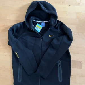 Svart NOCTA hoodie från Nike - Svart hoodie från Nike NOCTA med gul logga på bröstet och NOCTA-tryck på ärmen. Tröjan har huva, hel dragkedja och två fickor med dragkedja. Materialet är mjukt och känns som polyester. Snygga detaljer med broderade bokstäver på ryggen och ärmen.