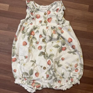 Romper med jordgubbar - Oanvänd och i nyskick. Supersöt romper från Newbie med jordgubbsmönster, storlek 56 (1-2mån). Volangdetaljer vid axlarna, träknappar framtill och resår och rynk vid benslut.