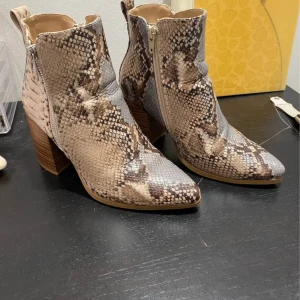 Beige boots med ormmönster och klack - Snygga boots med ormmönstrad yta i beige och bruna toner. Skorna har spetsig tå, grov träklack och dragkedja på sidan för enkel påtagning. Materialet är syntet med reptilstruktur som ger en cool vibe. Perfekt för dig som vill sticka ut med dina skor.