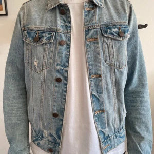 Ljusblå jeansjacka med slitningar från hollister - Snygg ljusblå jeansjacka med klassisk krage, bröstfickor med knappar och slitna detaljer för en cool vibe. Jackan har långa ärmar och bruna metallknappar framtill. Perfekt för dig som gillar en avslappnad och trendig stil.