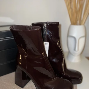 Mörkbruna lackstövletter med klack  - Snygga mörkbruna stövletter i glansigt lackmaterial med fyrkantig tå och kraftig klack. Dragkedja på insidan för enkel på- och avtagning. Perfekt för dig som vill sticka ut med ett trendigt statement. Perfekt till halloween om man vill klä ut sig till en bratz docka eller en 90/ tidigt 2000 tals icon! 