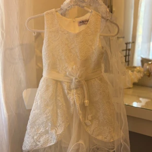 Vit spetsklänning Pretty Baby 6 år - Superfin vit klänning från Pretty Baby i spets med broderade detaljer och ett satinband i midjan med rosett. Ärmlös modell med rund hals och dragkedja bak. Ingår även en matchande slöja med blommor. Perfekt för speciella tillfällen!