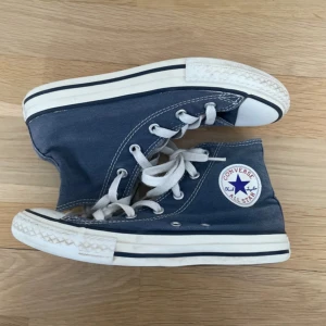 Blå Converse Chuck Taylor All Star - Säljer ett par klassiska Converse Chuck Taylor All Star sneakers i blått med vita snören och vit gummisula. Skorna har högt skaft, rund tå och tydliga vita sömmar. Ikonisk logga på sidan och platt sula. Perfekta för dig som gillar retrostil och streetwear.