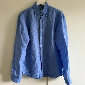 Blå linneskjorta Oscar Jacobson - Snygg blå skjorta från Oscar Jacobson i mjukt linne. Klassisk krage med knappar, vita knappdetaljer framtill och på manschetterna. Skjortan har lång ärm och en relaxed passform, perfekt för dig som gillar stilren och chill look.