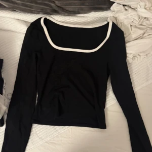 Svart långärmad topp med vit kant SHEIN - Svart långärmad topp från SHEIN med en snygg vit kant runt den fyrkantiga urringningen. Toppen är figurnära och har en stilren, minimalistisk look som passar perfekt till jeans eller kjol. Materialet är mjukt och stretchigt för en bekväm känsla.