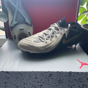 Nike fotbollsskor svart/vit/guld - Säljer ett par Nike fotbollsskor i svart, vitt och guld. Skorna har snörning, platt sula med dobbar och en snygg guldig swoosh på sidan. Ovandelen är i syntetmaterial och designen är modern och sportig – perfekta för dig som vill sticka ut på planen.