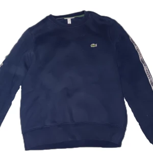 Marinblå sweatshirt från Lacoste - Säljer en marinblå sweatshirt från Lacoste med klassisk rund hals och långa ärmar. Tröjan har vita logoband längs ärmarna och den ikoniska gröna krokodilen broderad på bröstet. Nypris 1500, tveka inte att höra av er vid frågor