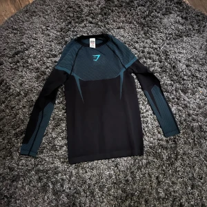Svart långärmad träningströja Gymshark - Svart och mörkblå långärmad träningströja från Gymshark med sportig design och logga på bröstet. Tillverkad i stretchigt material med paneler i mesh-liknande struktur för extra ventilation. Perfekt för gymmet eller löprundan . Säljer den eftersom den inte passade helt oanvänd 