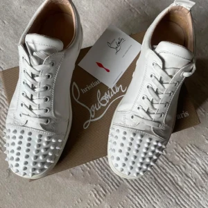 Christian Louboutin vita sneakers med nitar - Säljer ett par vita sneakers från Christian Louboutin i skinn med ikoniska röda sulor och coola nitar på tån. Skorna har snörning och en klassisk låg siluett. Perfekta för dig som vill sticka ut med en lyxig och edgy vibe.