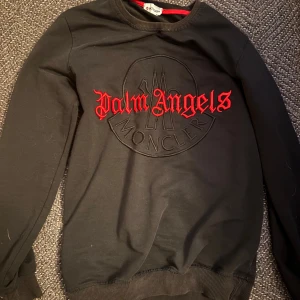 Svart Palm Angels x Moncler sweatshirt - Svart sweatshirt från Palm Angels x Moncler med broderad logga och röd Palm Angels-text på bröstet. Tröjan har rund hals, ribbade muddar och är tillverkad i mjuk bomull. Perfekt för dig som gillar streetwear och exklusiva samarbeten.