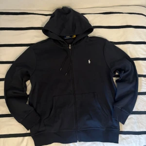 Mörkblå hoodie från Polo Ralph Lauren - Snygg mörkblå hoodie från Polo Ralph Lauren med klassisk broderad logga på bröstet. Tröjan har huva med snörning, dragkedja framtill och två stora fickor. Tillverkad i mjuk bomull, perfekt för chill dagar. Passformen är normal och ärmarna är långa. Hör av dig om du har några frågor.
