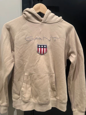 Beige hoodie från Gant med brodyr - Snygg beige hoodie från Gant med broderad logga och sköld på bröstet. Tröjan har huva, känguruficka och är tillverkad i mjuk bomull. Perfekt för en chill och stilren look.