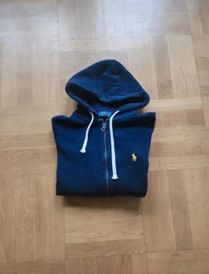 Mörkblå hoodie från Polo Ralph Lauren - Mörkblå hoodie från Polo Ralph Lauren med dragkedja, vit snörning i huvan och klassisk gul logga på bröstet. Tröjan är i mjuk bomull och har långa ärmar samt ribbade muddar. Perfekt för en chill och stilren look.