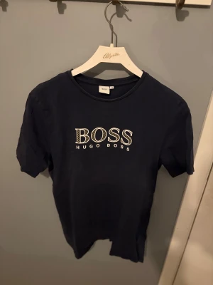 Mörkblå Hugo Boss t-shirt - Snygg mörkblå t-shirt från Hugo Boss med stort BOSS-tryck i vitt på bröstet. Klassisk rund hals och korta ärmar. Tillverkad i mjuk bomull som känns skön mot huden. Perfekt för dig som gillar stilrena och trendiga märkesplagg.