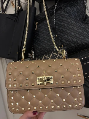 Beige axelväska med nitar - Snygg beige axelväska i quiltad design med guldiga nitar och kedjerem. Storlek är rymlig och bra. Den är i bra skick och är aldrig använd. Tveka inte på att fråga om du undrar något💕