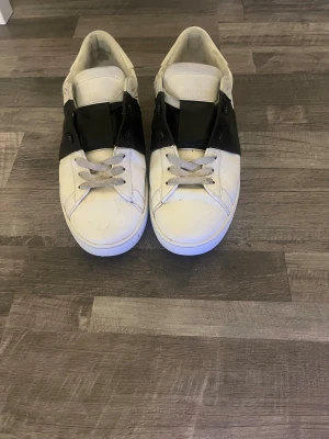 Valentino Garavani open vita sneakers - Snygga vita sneakers från Valentino Garavani med bred svart läderdetalj över snörningen. Klassisk rund tå och platt sula, med coola nitar längs hälen. Skorna har snörning och är tillverkade i skinn för en lyxig känsla.