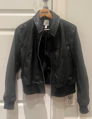 Svart skinnjacka från H&M - Snygg svart skinnjacka från H&M i storlek S. Jackan har klassisk krage, ribbade muddar och nederkant samt dragkedja framtill. Perfekt för dig som gillar en stilren look med lite edge. Materialet är syntetiskt skinn med mjuk insida.