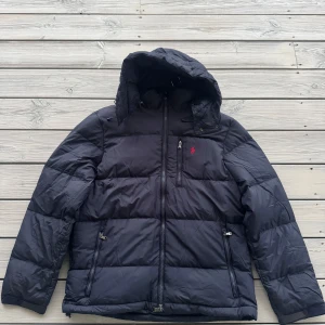 Marinblå dunjacka Ralph Lauren - Marinblå pufferjacka från Polo Ralph Lauren med huva, flera dragkedjefickor och klassisk röd Polo-logga på bröstet. Jackan är lite tillplattad eftersom den hängt tillsammans med andra jackor. Men kommer puffas upp till normalt läge vid användning.